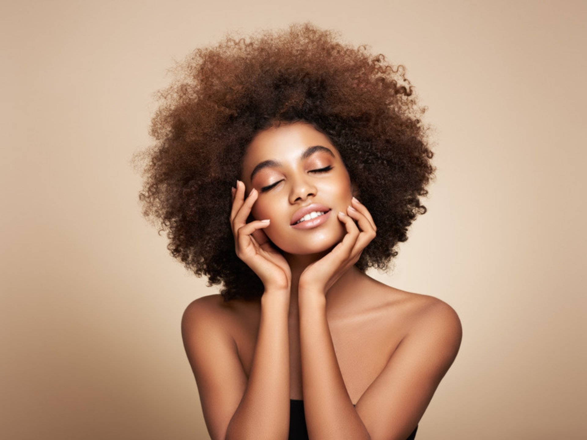 Woman with a natural afro smiling with her eyes closed and resting her hands on her face